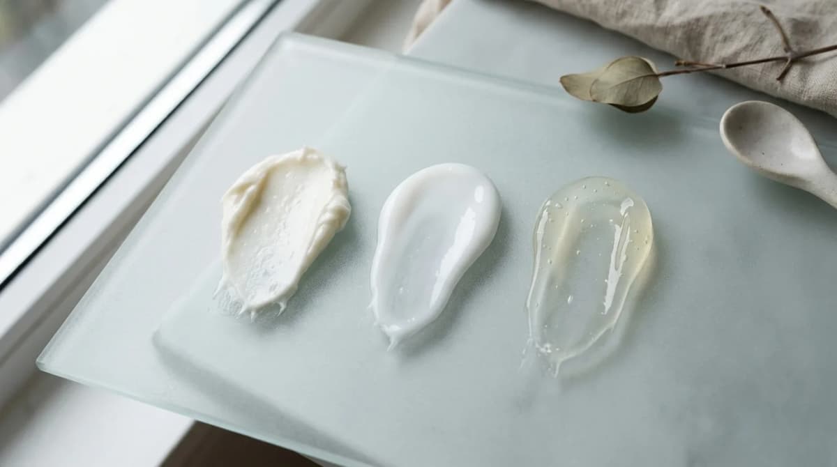 Flat lay beauty editorial photography of three cosmetic cream textures on frosted glass surface — a thick opaque balm, a lightweight milky lotion, and a translucent gel — showing different consistencies side by side, natural daylight, minimalist styling